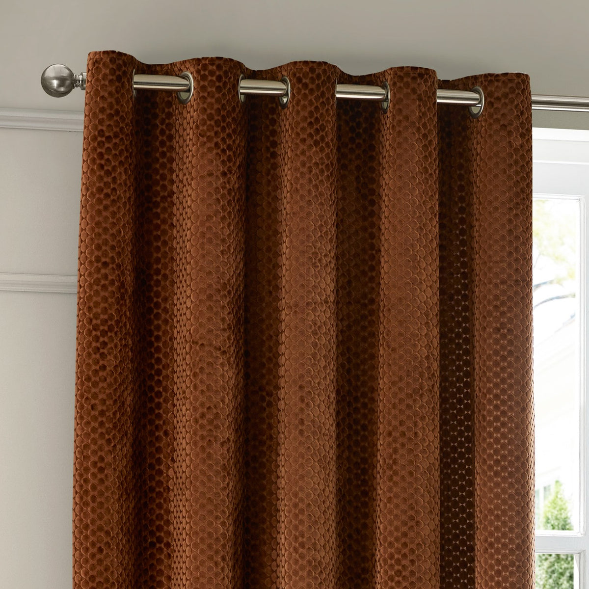 Neptune Velvet Fully Lined Eyelet Curtains in copper, showing honeycomb-textured velvet fabric on silver eyelet rings and pole, hung beside a window.