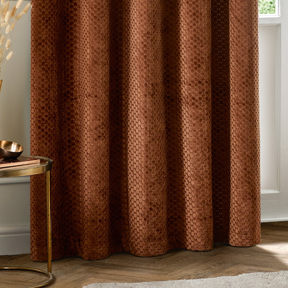 Neptune Velvet Fully Lined Eyelet Curtains in copper, showing the honeycomb-textured fabric in soft folds, styled in a living room beside a round brass side table.