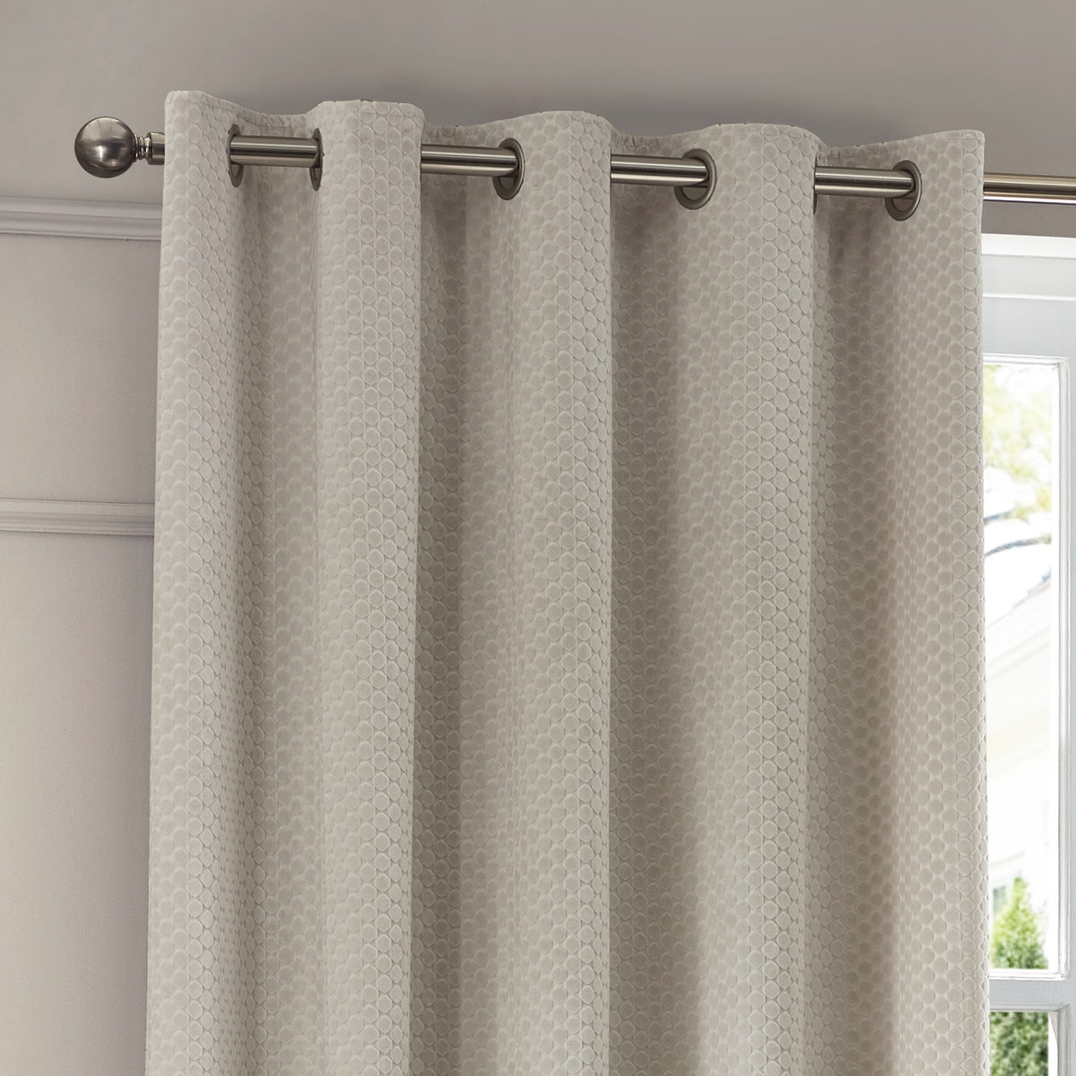 Neptune Velvet Fully Lined Eyelet Curtains in ivory with geometric honeycomb pile, shown hung on a brushed metal pole beside a window.