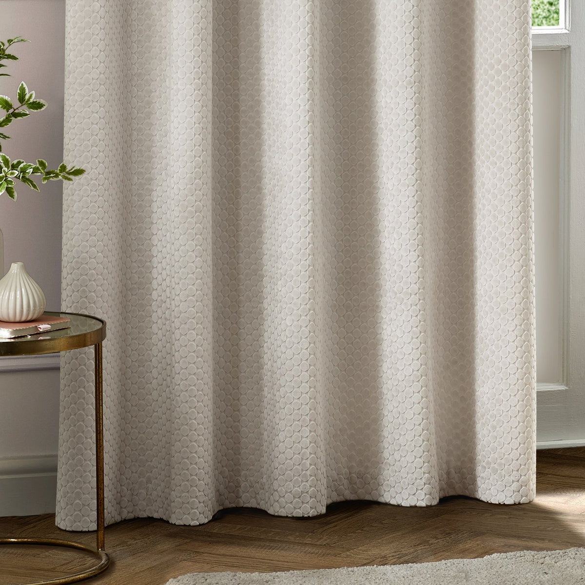 Neptune Velvet Fully Lined Eyelet Curtains in ivory, showing a honeycomb dot cut-pile texture, styled in a living room beside a round side table and potted plant.