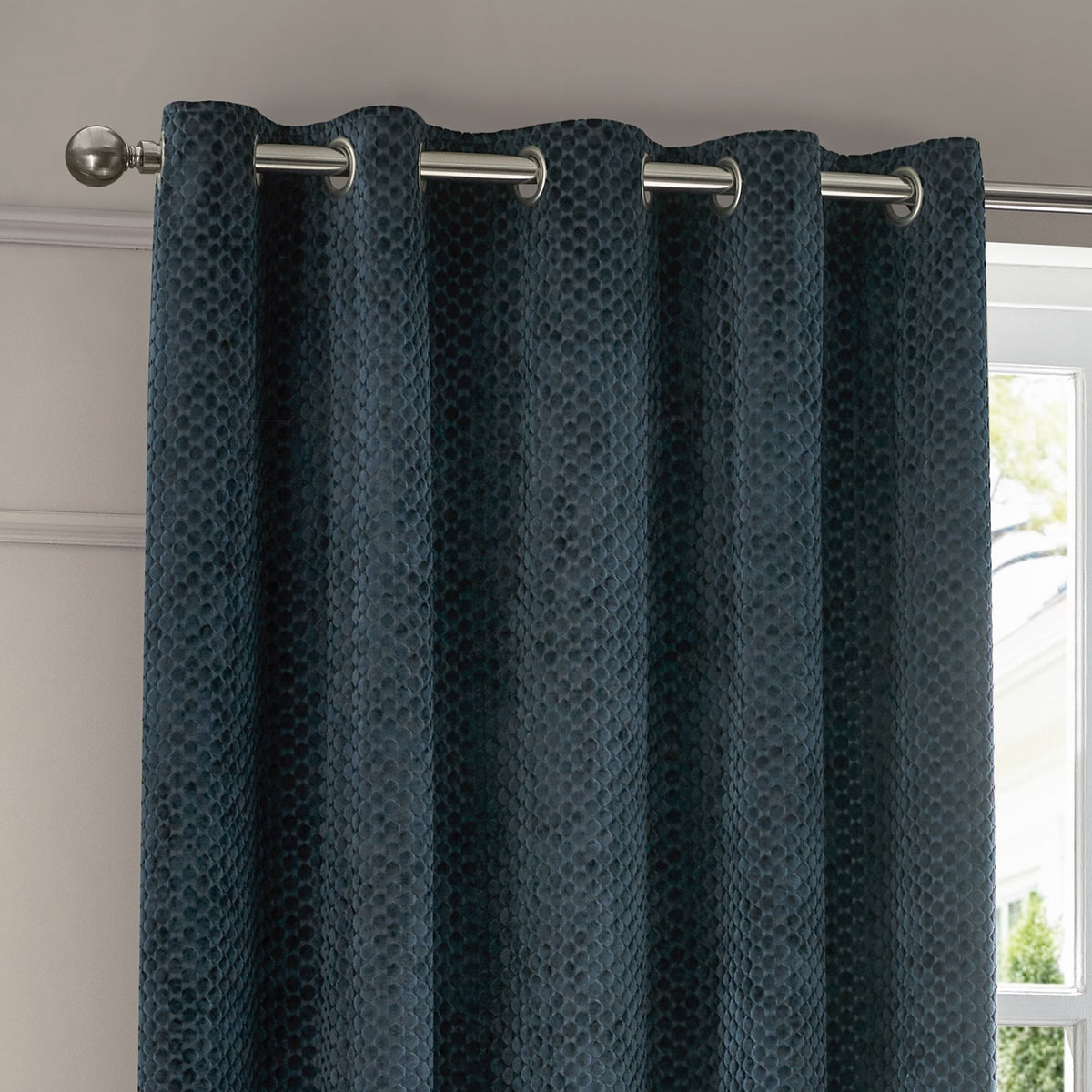 Neptune Velvet Fully Lined Eyelet Curtains in navy, showing textured cut-velvet fabric with dotted geometric pattern hung on a chrome eyelet rail beside a window.