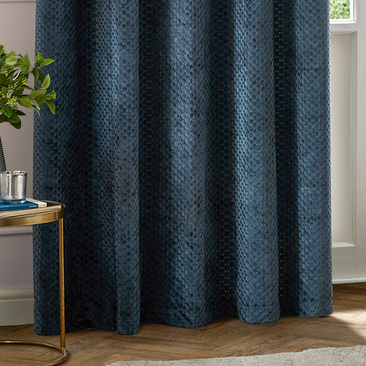 Neptune Velvet Fully Lined Eyelet Curtains in navy, shown with eyelet heading and textured geometric pile, hanging in a living room beside a wooden side table and plant.