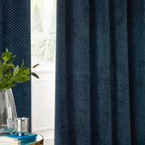 Navy velvet eyelet curtains with a raised geometric dot pattern, shown fully lined and gathered beside a window in a living room setting.