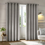 Neptune Velvet Fully Lined Eyelet Curtains in silver grey with a geometric textured pile, hung on a chrome eyelet rod beside a glass door in a living room setting.