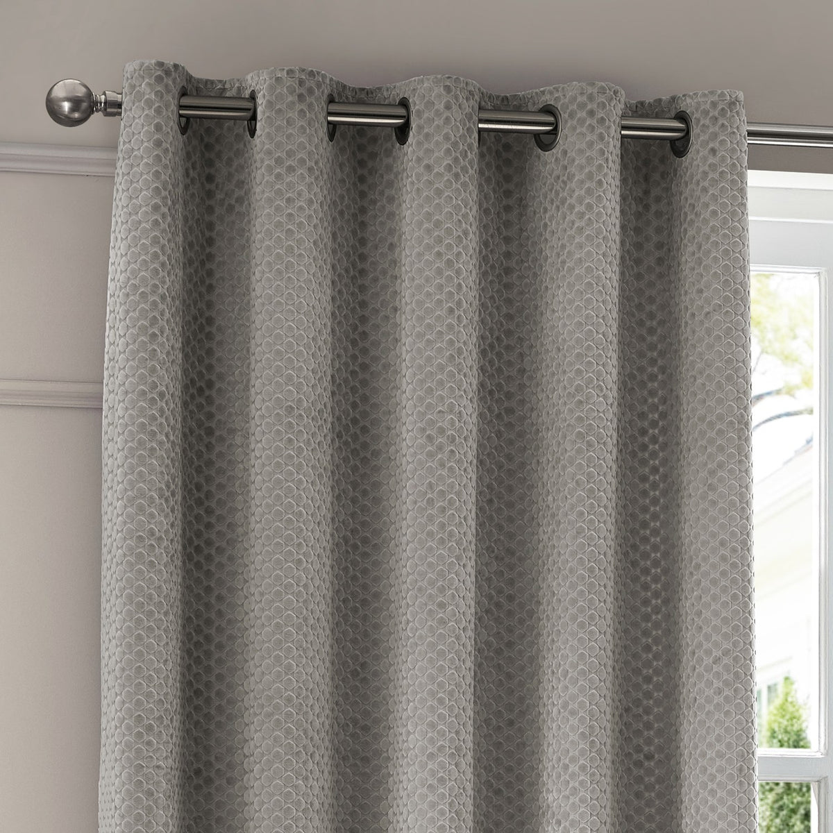 Neptune Velvet Fully Lined Eyelet Curtains in silver grey with geometric cut-velvet pattern, eyelet heading on a chrome pole in a living room setting.