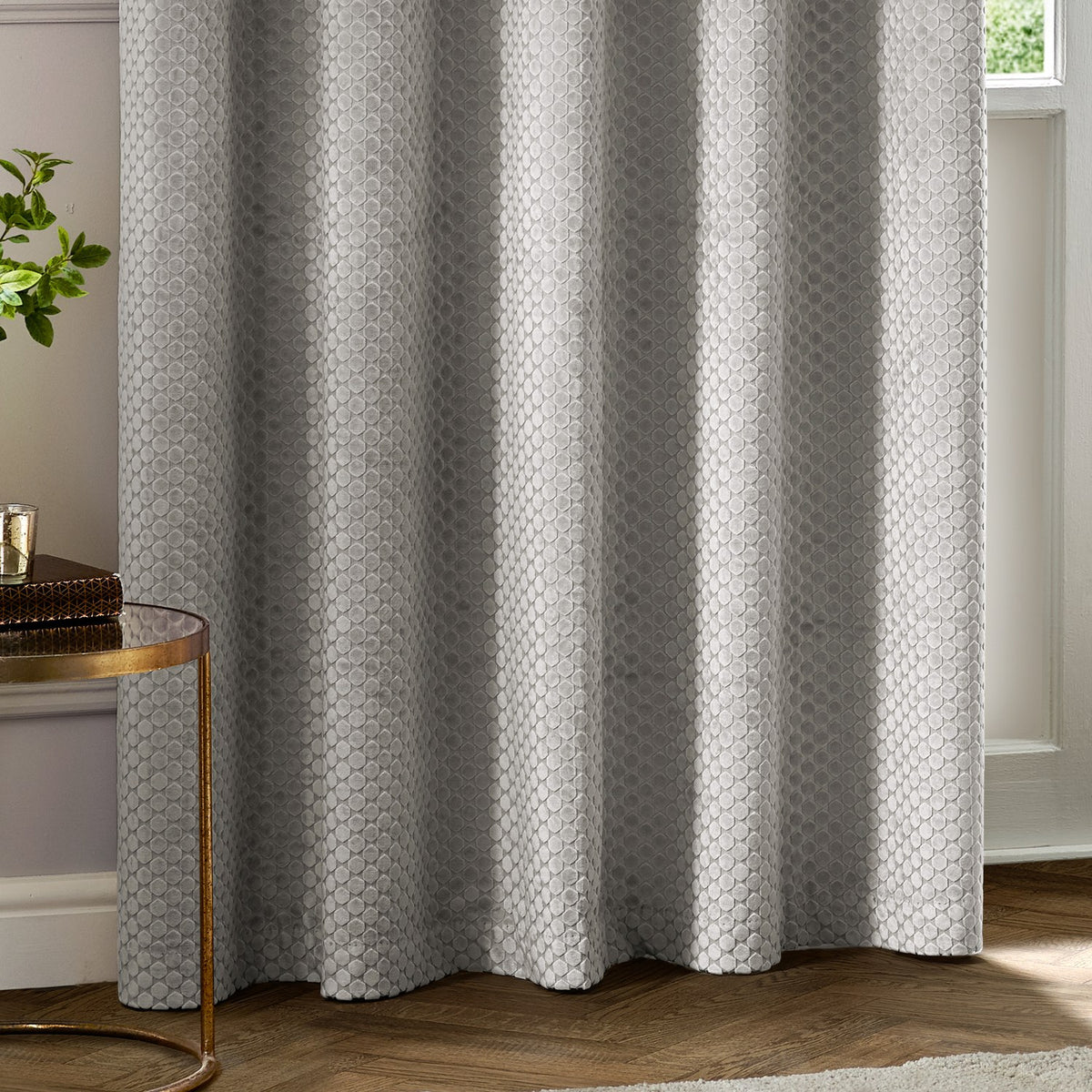 Neptune Velvet Fully Lined Eyelet Curtains in silver grey with a geometric cut-pile pattern, shown draped in a living room beside a round side table and window.