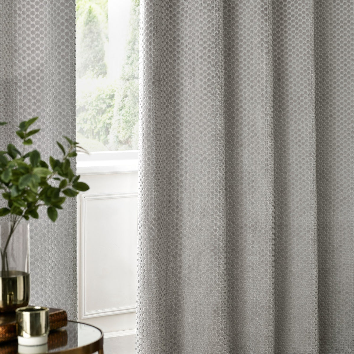 Neptune Velvet Fully Lined Eyelet Curtains in silver grey with geometric cut-pile texture, shown hanging in eyelet folds beside a window in a living room setting.