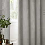 Neptune Velvet Fully Lined Eyelet Curtains in silver grey with geometric cut-pile texture, shown hanging in eyelet folds beside a window in a living room setting.