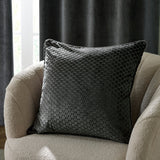 Neptune Geometric Velvet Cushion in charcoal, square with raised honeycomb pattern, shown on a cream boucle armchair against dark textured curtains.