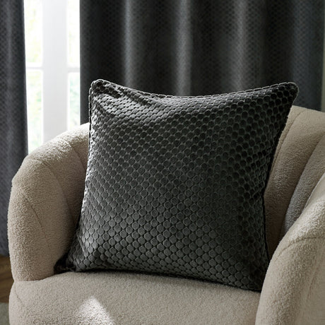 Neptune Geometric Velvet Cushion in charcoal, square with raised honeycomb pattern, shown on a cream boucle armchair against dark textured curtains.