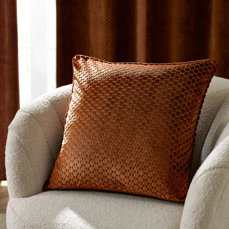 Neptune Geometric Velvet Cushion in copper orange with a honeycomb textured pattern, shown on a cream bouclé armchair against a dark curtain backdrop.