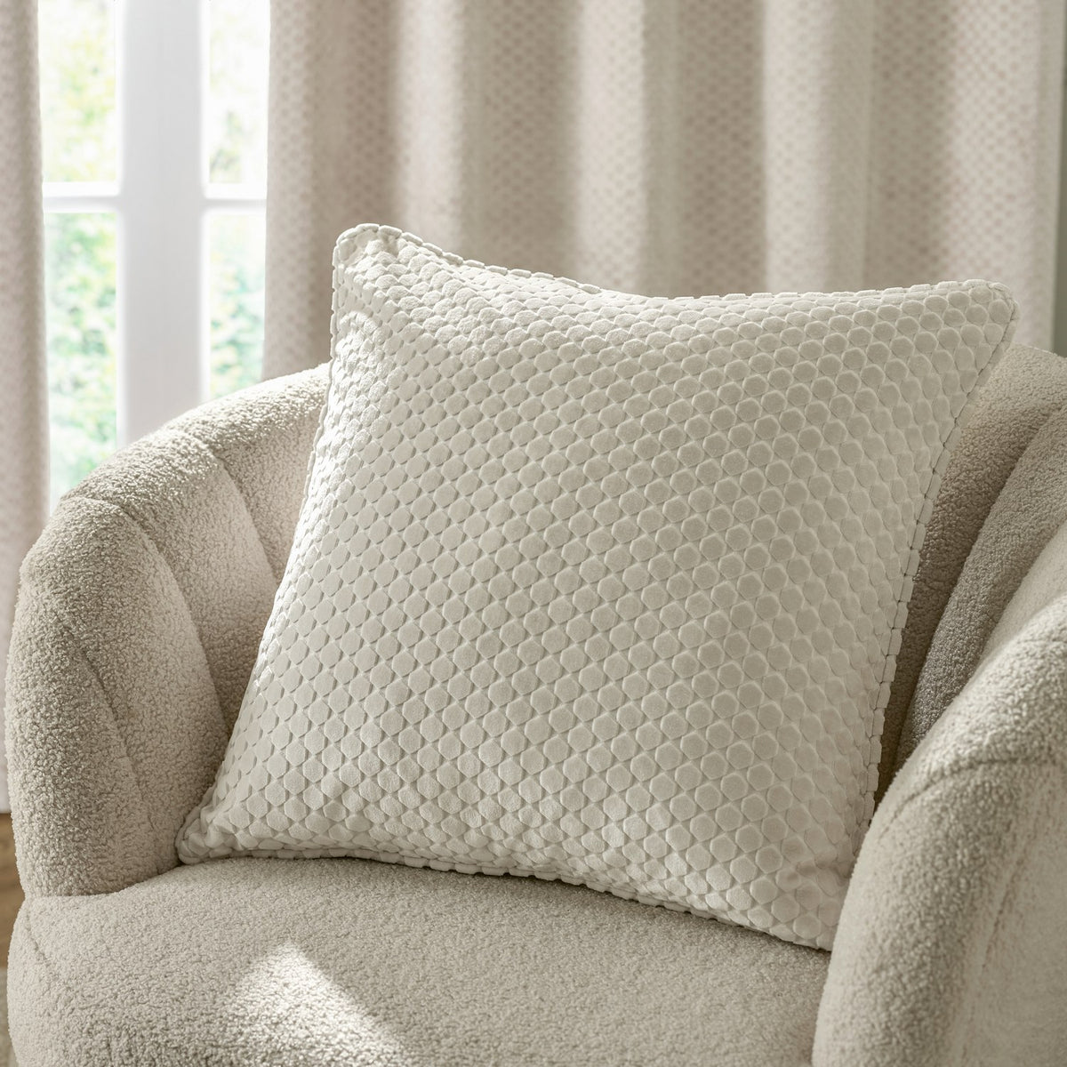 Neptune Geometric Velvet Cushion in ivory with a raised honeycomb-style pattern, shown on a boucle armchair beside a curtained window.