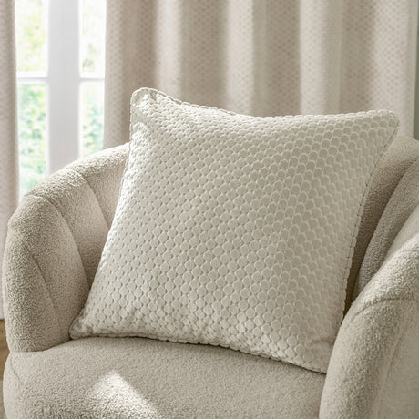 Neptune Geometric Velvet Cushion in ivory with a raised honeycomb-style pattern, shown on a boucle armchair beside a curtained window.
