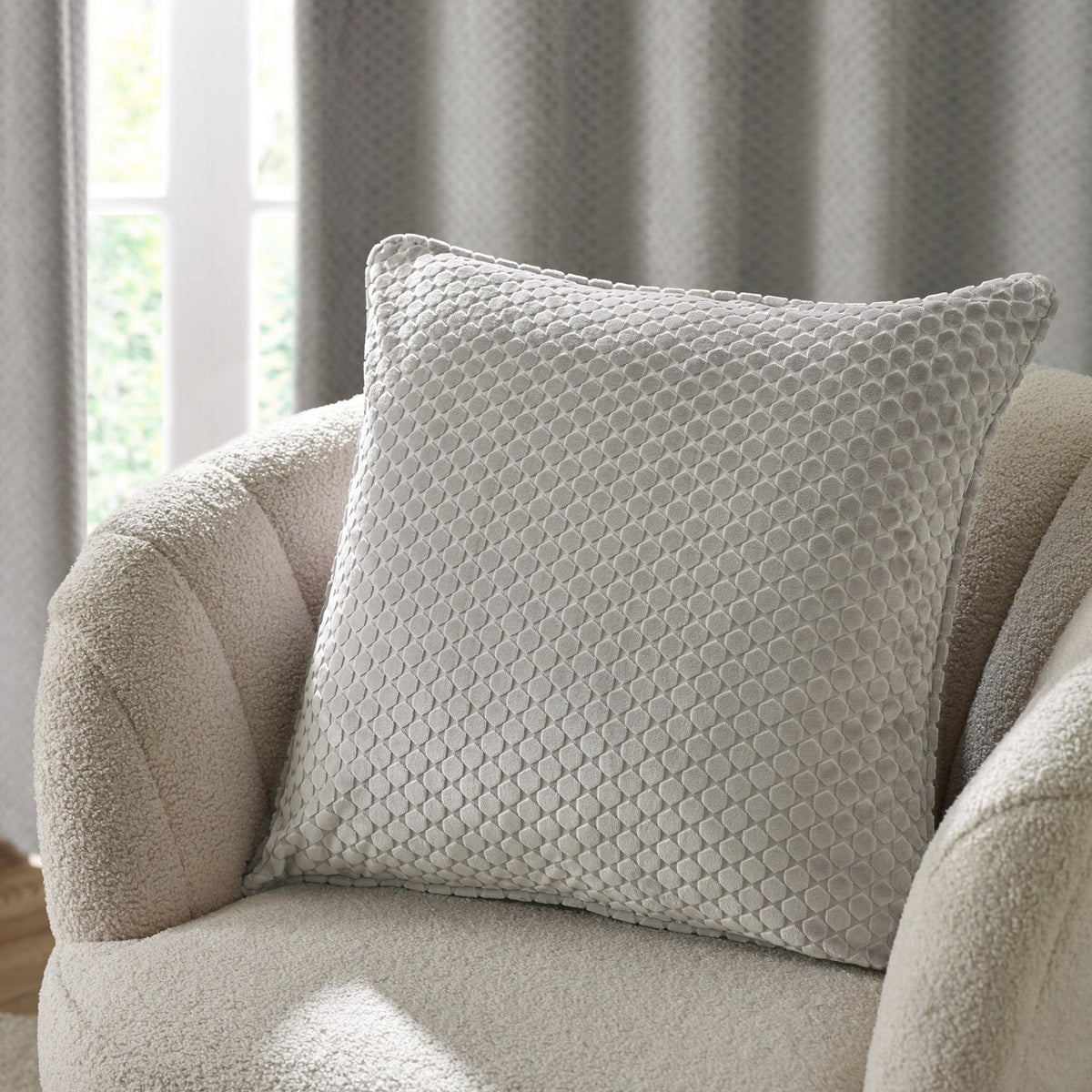 Neptune Geometric Velvet Cushion in silver grey with raised honeycomb dot texture, shown on a cream boucle armchair against curtains.