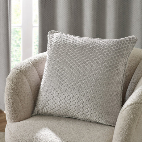 Neptune Geometric Velvet Cushion in silver grey with raised honeycomb dot texture, shown on a cream boucle armchair against curtains.