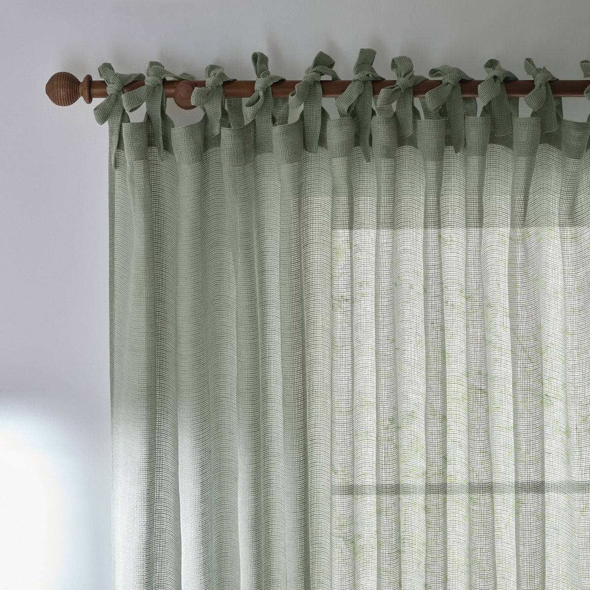 Nimbus sheer tie-top curtain panel in eucalyptus green polyester, fabric ties knotted over a wooden pole, showing the woven texture against a light room.