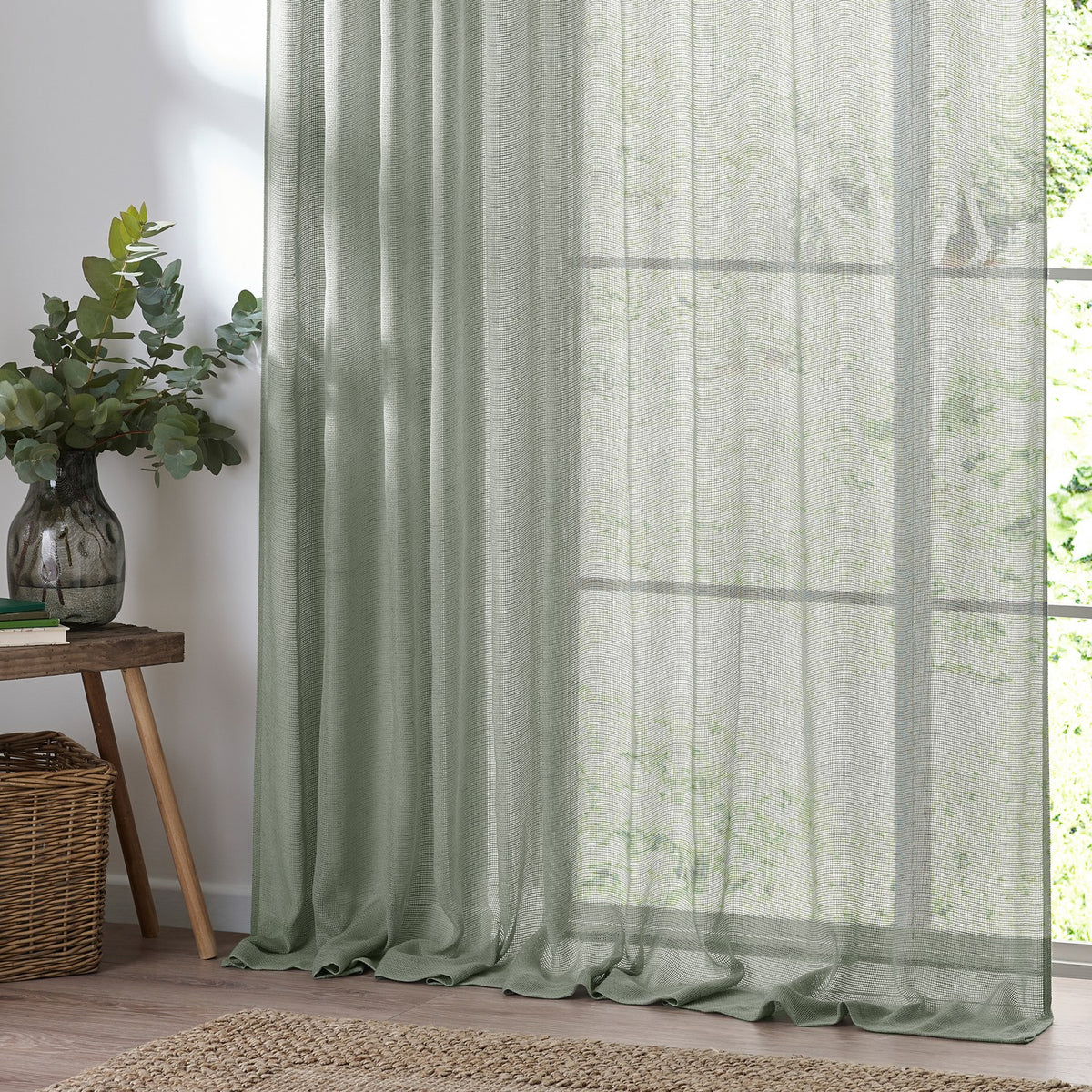 Nimbus sheer tie-top curtain panel in eucalyptus green polyester, hung by a bright window beside a plant-topped side table in a living room.