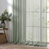 Nimbus sheer tie-top curtain panel in eucalyptus green polyester, hung by a bright window beside a plant-topped side table in a living room.