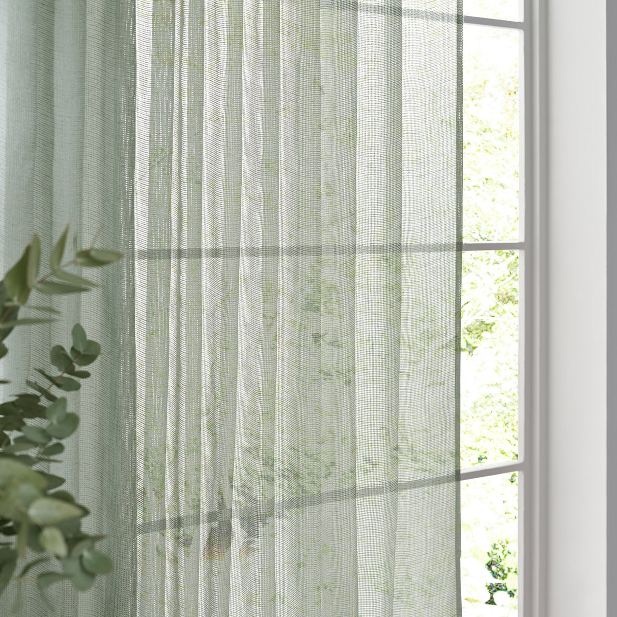 Nimbus sheer tie-top curtain panel in eucalyptus green polyester, hanging by a window with soft daylight and greenery visible through the fabric.