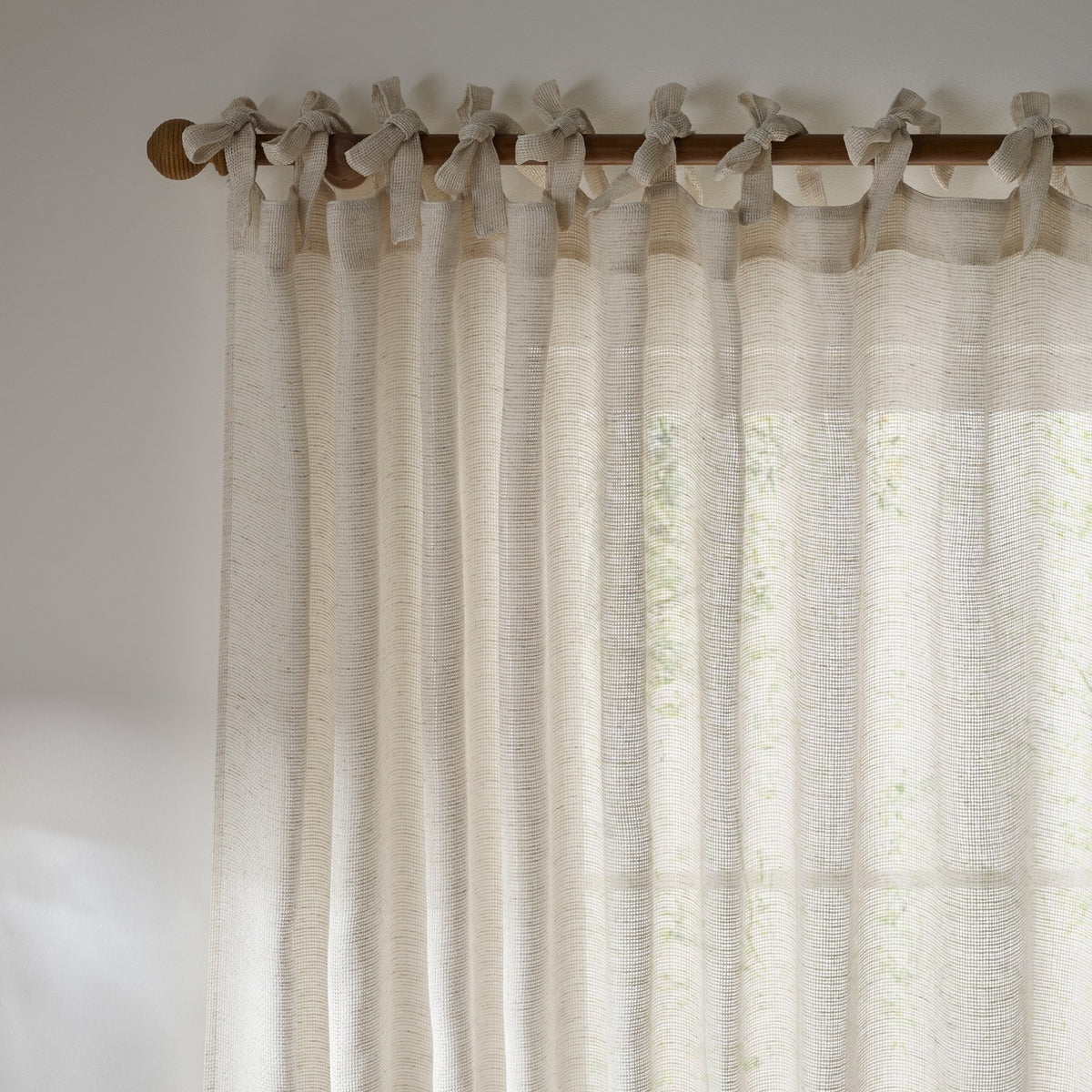 Nimbus sheer tie-top curtain panel in natural polyester, hung from a wooden pole with fabric tie knots, showing a loosely woven, light-filtering texture against a window.