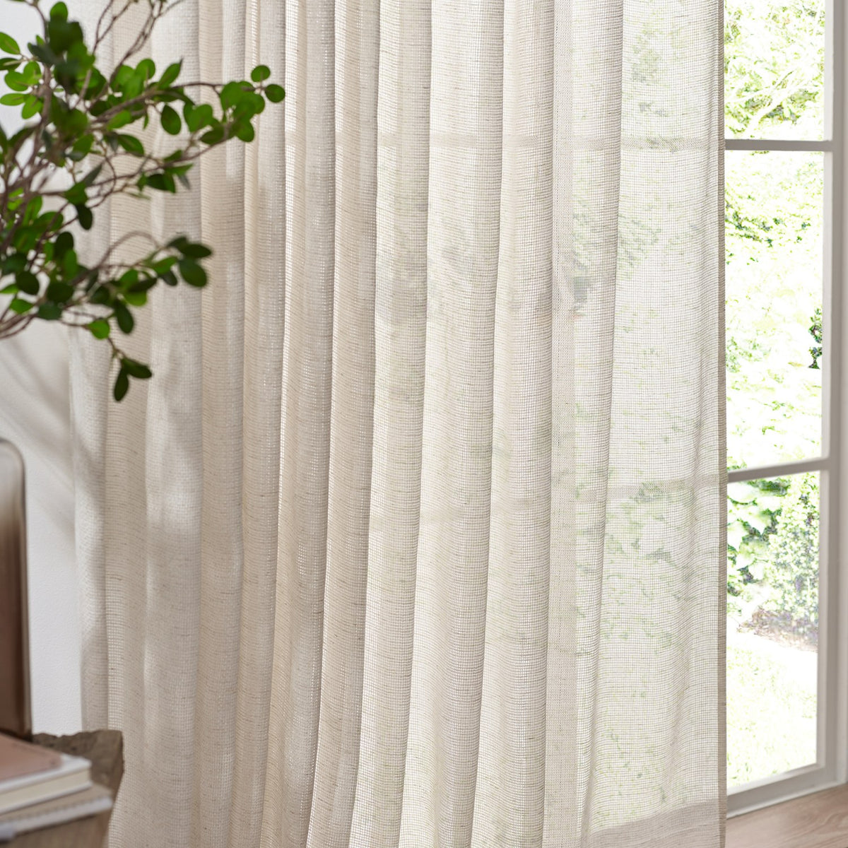 Nimbus sheer polyester curtain panel in natural, shown pleated at a window beside a green plant, its loose weave softly filtering daylight.