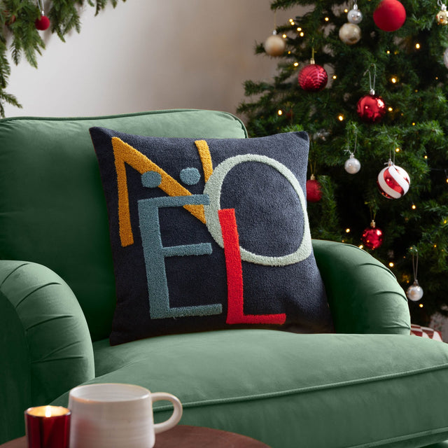 Noel Plain Polyester Cushion in charcoal-navy with tufted NOEL lettering in amber, blue, grey-green and red, shown on a green armchair beside a Christmas tree.