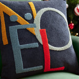 Noel Plain Polyester Cushion in charcoal-navy with tufted "NOEL" lettering in amber, blue, grey-green and red, shown on a green sofa.