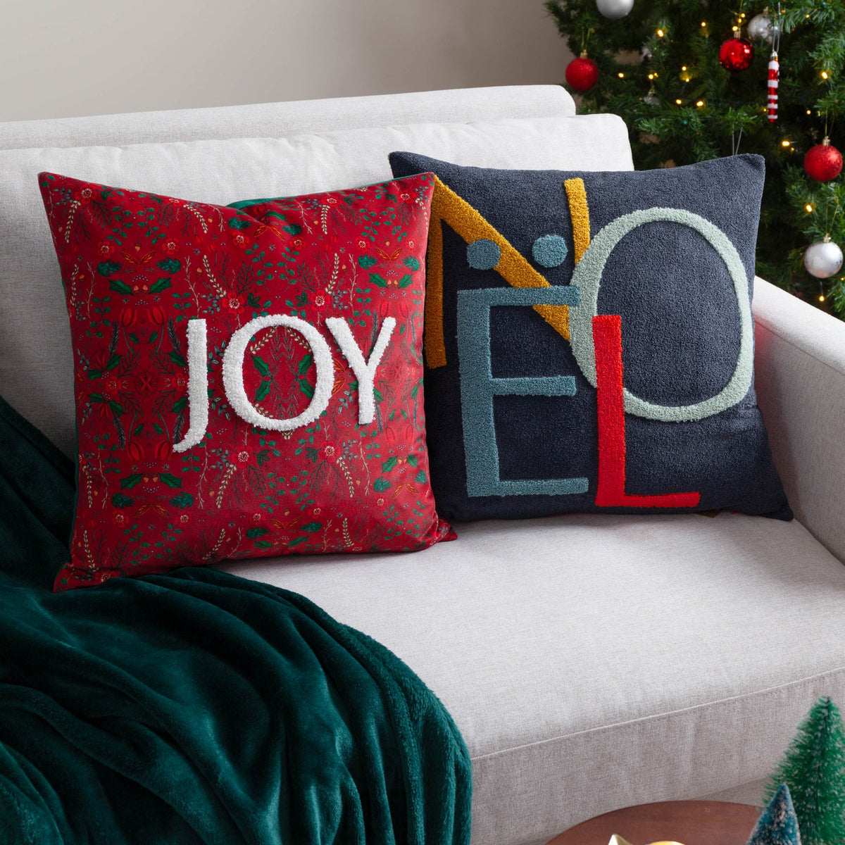 Noel Plain Polyester Cushion in charcoal-navy with tufted NOEL lettering, shown beside a red floral JOY cushion on a sofa near a Christmas tree.