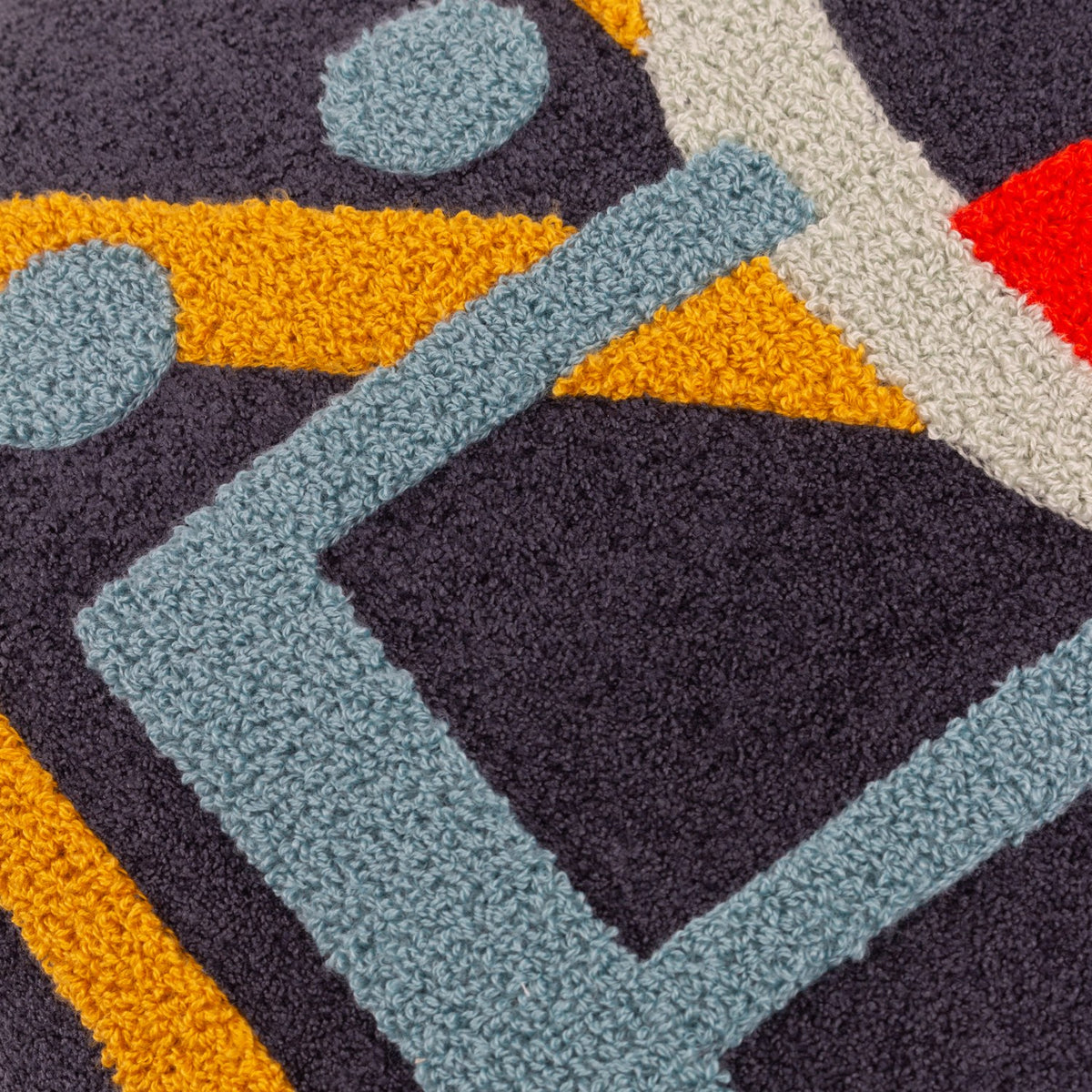 Close-up of the Noel Plain Polyester Cushion in charcoal-navy, showing tufted loop-pile lettering in amber, pale blue, grey-green and red forming part of the word NOEL.