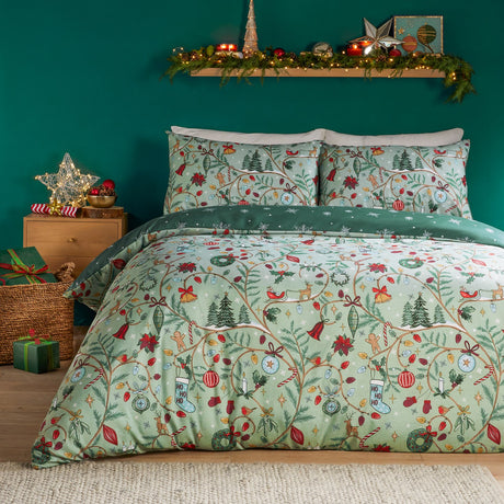 Noelle Printed Reversible Duvet Cover Set in green, shown on a bedroom bed with festive Christmas trees, baubles, bells and gingerbread motifs on a soft green ground, matching pillowcases included.