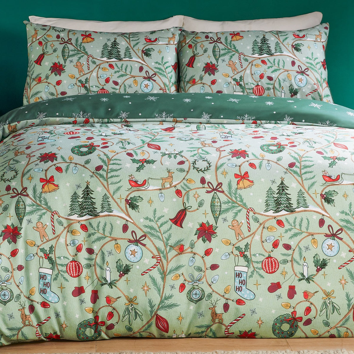 Noelle Printed Reversible Duvet Cover Set in green, styled on a bed with matching pillowcases showing a festive print of trees, wreaths, baubles and robins against a soft green ground.