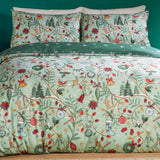 Noelle Printed Reversible Duvet Cover Set in green, styled on a bed with matching pillowcases showing a festive print of trees, wreaths, baubles and robins against a soft green ground.