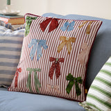 Noortje Velvet Silk Cushion in Silk Pink on a blue sofa, featuring multicoloured bow prints on a fine red and white striped velvet face with red piped edging.