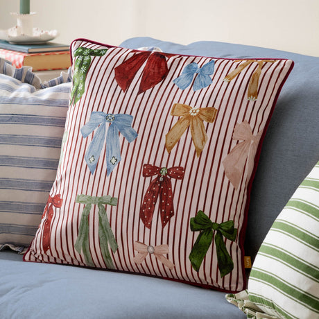 Noortje Velvet Silk Cushion in Silk Pink on a blue sofa, featuring multicoloured bow prints on a fine red and white striped velvet face with red piped edging.
