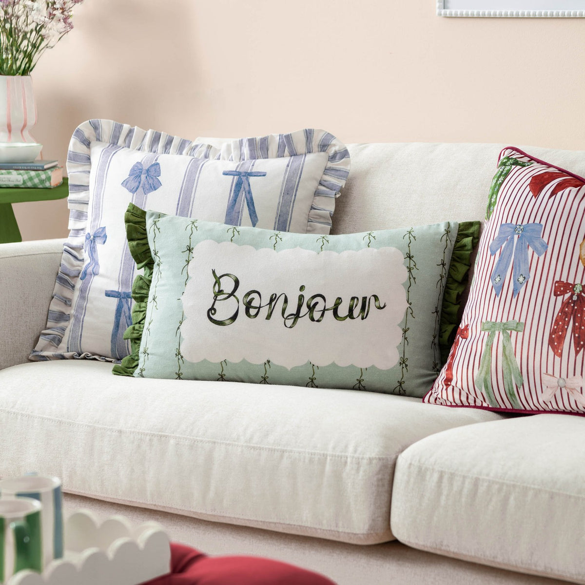 Bonjour lumbar cushion styled on a cream sofa with striped ribbon-print and pink bow-patterned scatter cushions in a living room setting.