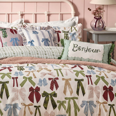 Noortje Silk Abstract Duvet Cover Set in pink polycotton, printed with bows in green, blue, red and gold, styled on a bed with a "Bonjour" cushion in a pink bedroom.