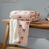 Noortje Bows Silk Throw in Silk Pink, folded on a wooden bench with sherpa fleece reverse showing, printed with red bow and stripe pattern on a white ground.