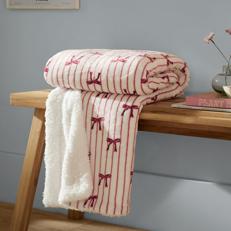 Noortje Bows Silk Throw in Silk Pink, folded on a wooden bench with sherpa fleece reverse showing, printed with red bow and stripe pattern on a white ground.