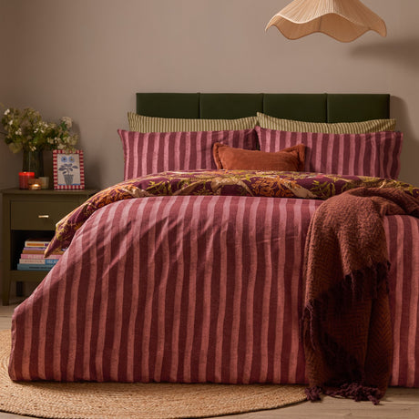 Nya Woodland Reversible Duvet Cover Set in burgundy, showing the striped rusty pink and plum side, styled on a bed with a green headboard and rust cushions.