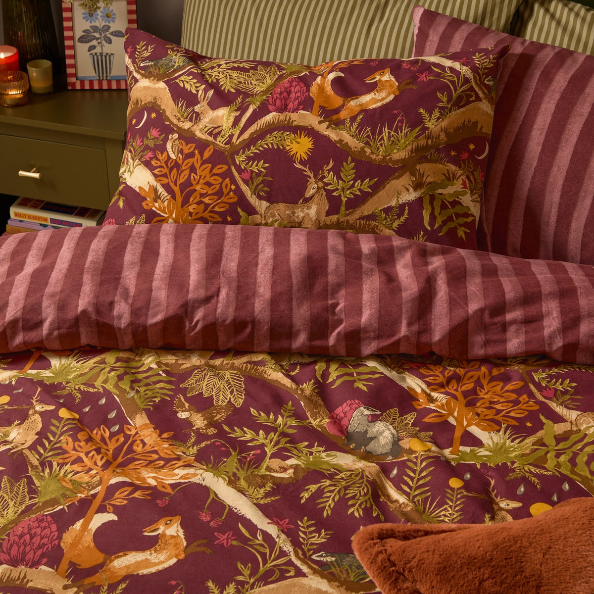 Nya Woodland Reversible Striped Duvet Cover Set in burgundy on a bed, showing the woodland animal print side and reverse pink and plum striped fabric with matching pillowcase.