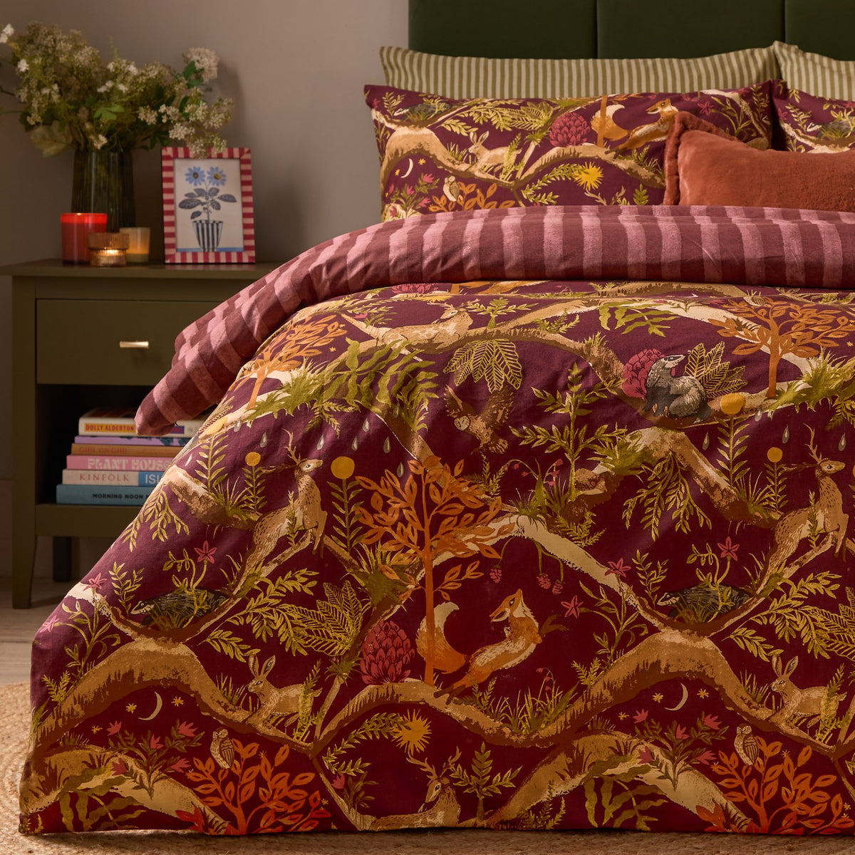 Nya Woodland Reversible Duvet Cover Set in burgundy, featuring a woodland animal print with foxes, hares and owls, styled on a bed with striped reverse edge visible in a bedroom setting.