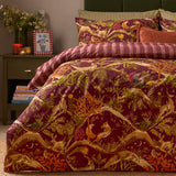 Nya Woodland Reversible Duvet Cover Set in burgundy, featuring a woodland animal print with foxes, hares and owls, styled on a bed with striped reverse edge visible in a bedroom setting.