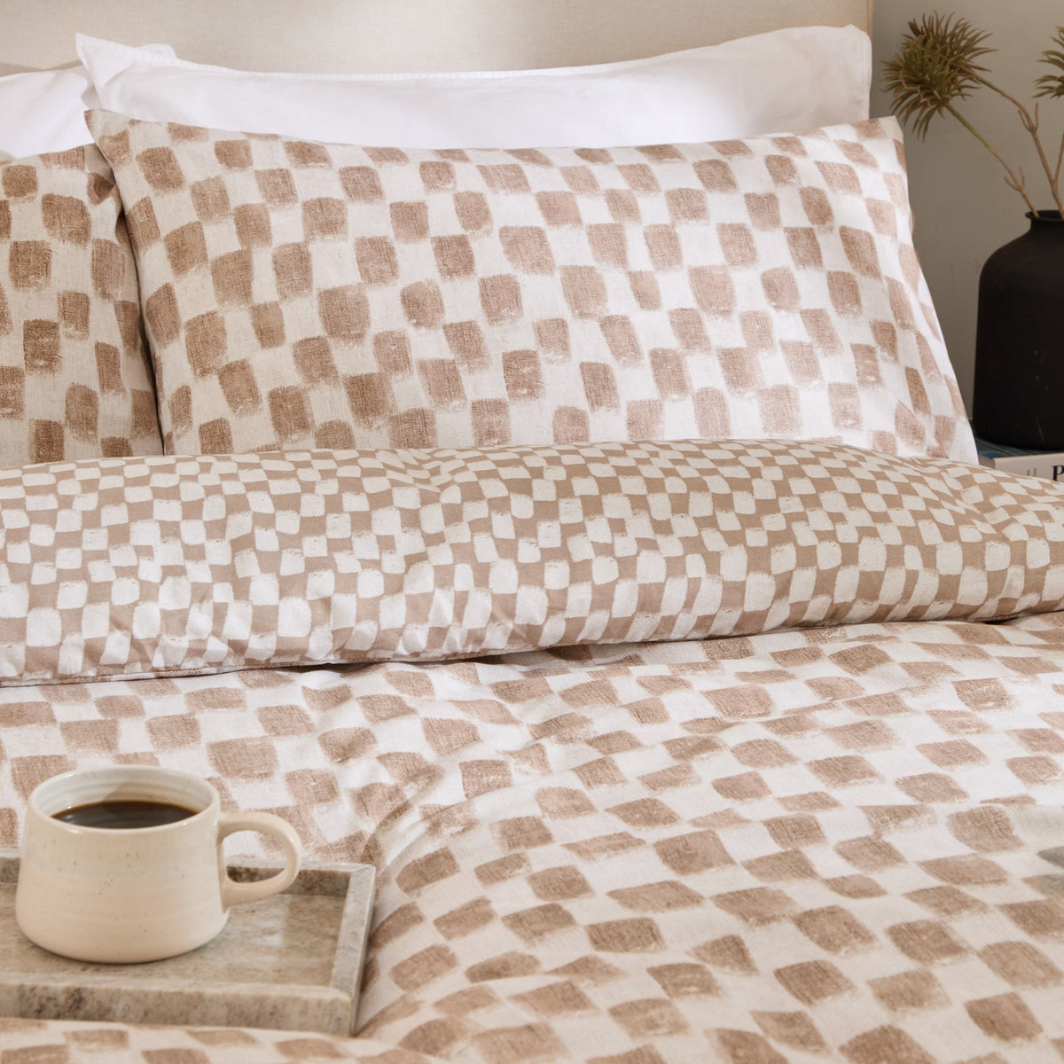 Nyhavn Reversible Check Duvet Cover Set in Tobacco, shown on a bed with matching pillowcases in a tan and off-white checkerboard pattern, styled in a bedroom with a coffee mug on a tray.