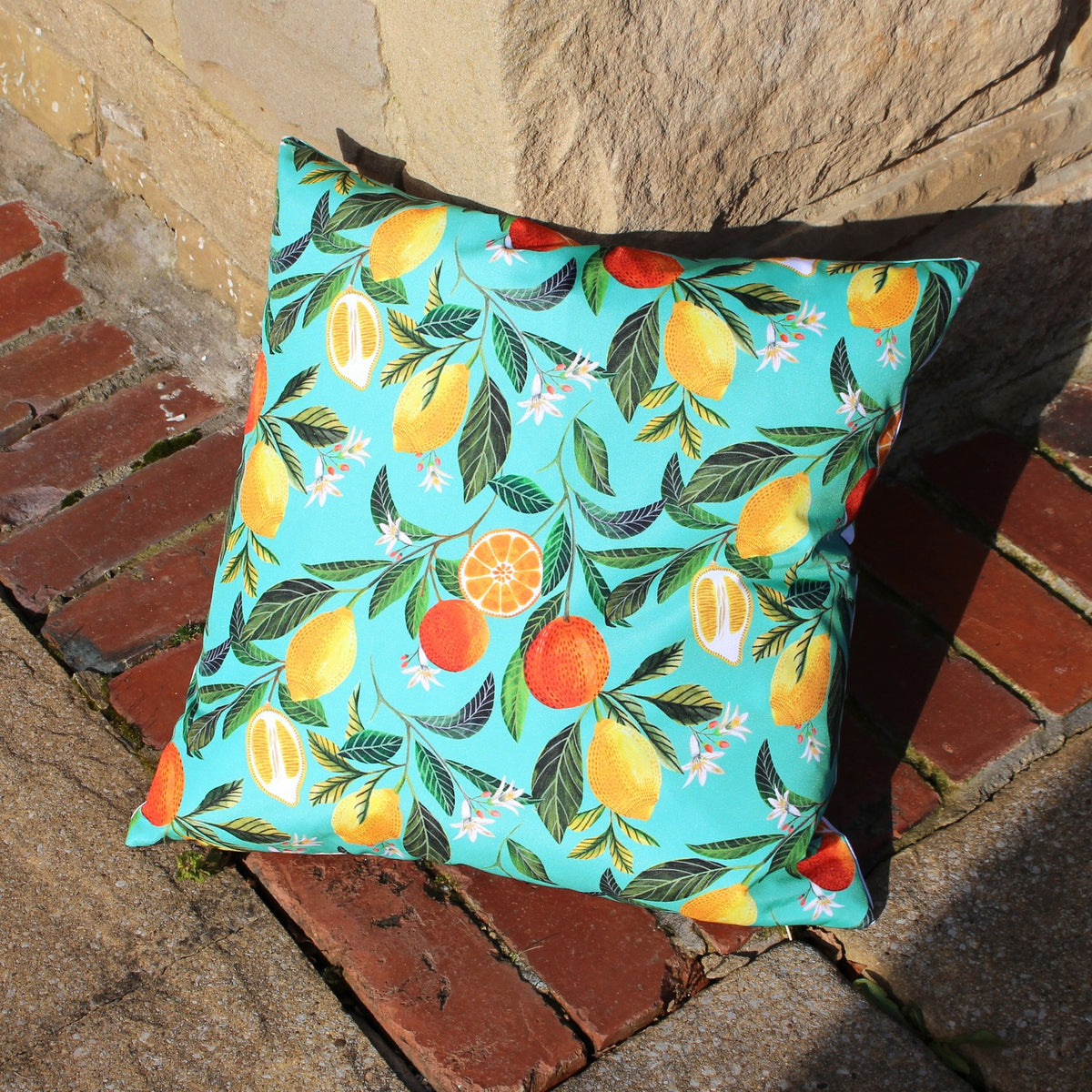 Square Orange Blossom outdoor cushion in polyester with a turquoise-blue print of lemons, oranges and leaves, shown outdoors on a brick and stone step.