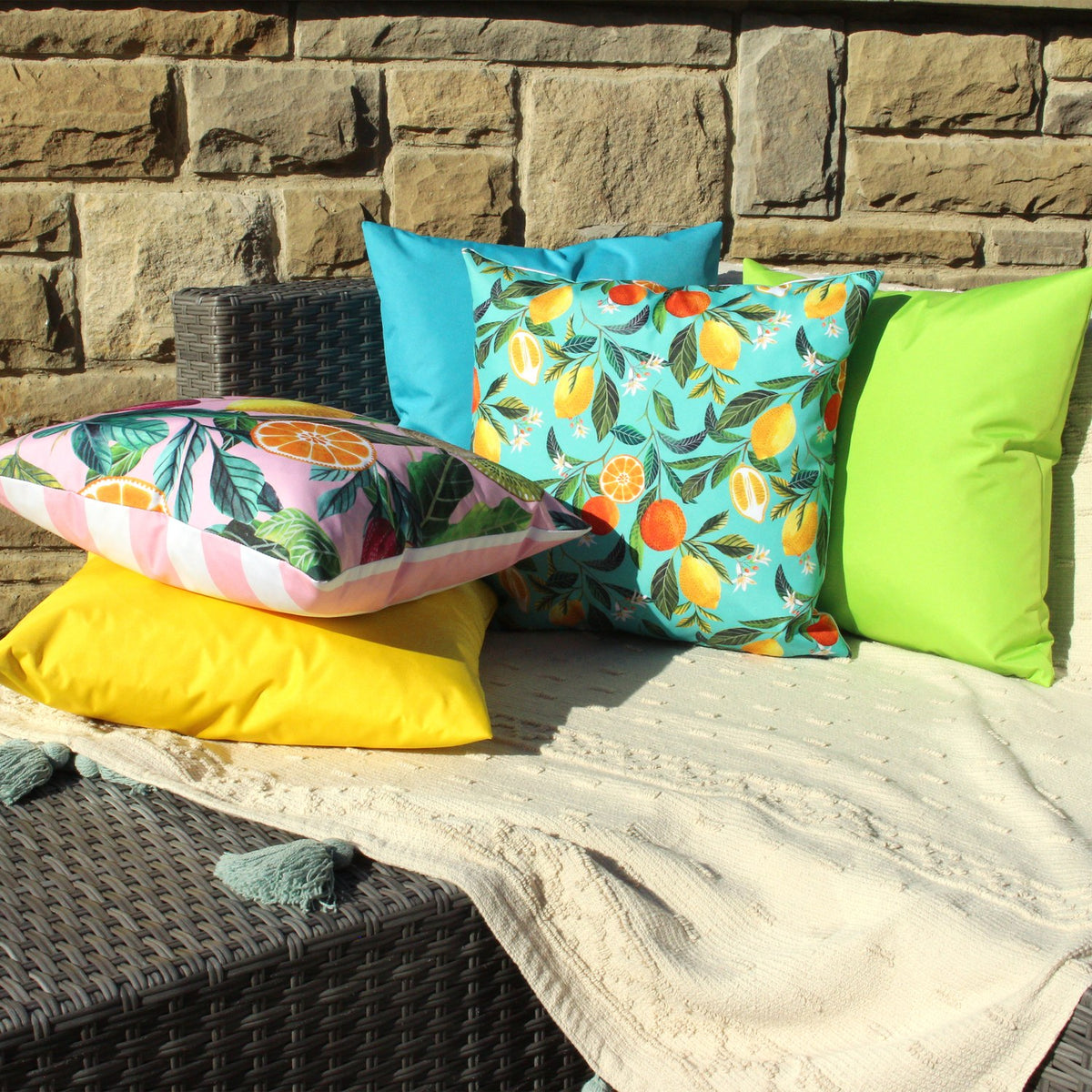 Orange Blossom Abstract polyester cushion in turquoise with citrus print, styled outdoors alongside yellow, pink-striped, teal and green cushions on a wicker bench with a cream throw.