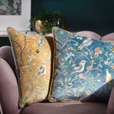 Orient Animal Velvet Cushion in gold with hand-painted bird and floral design, shown on a pink sofa beside a matching teal cushion in a living room setting.