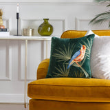 Odisha Exotic Bird Animal Velvet Cushion in emerald green with gold embroidered fronds, shown on a mustard velvet sofa in a living room setting.