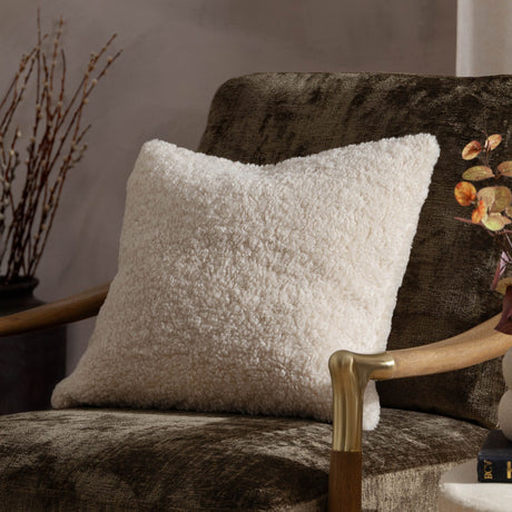 Olann Velvet Faux Fur Cushion in natural beige, square shape with dense curly shearling-look pile, shown on a brown velvet armchair.