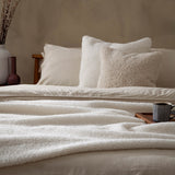 Olann Velvet Faux Fur Cushion in natural beige with curly shearling-look pile, styled on a bed with linen bedding and a chunky knit throw.