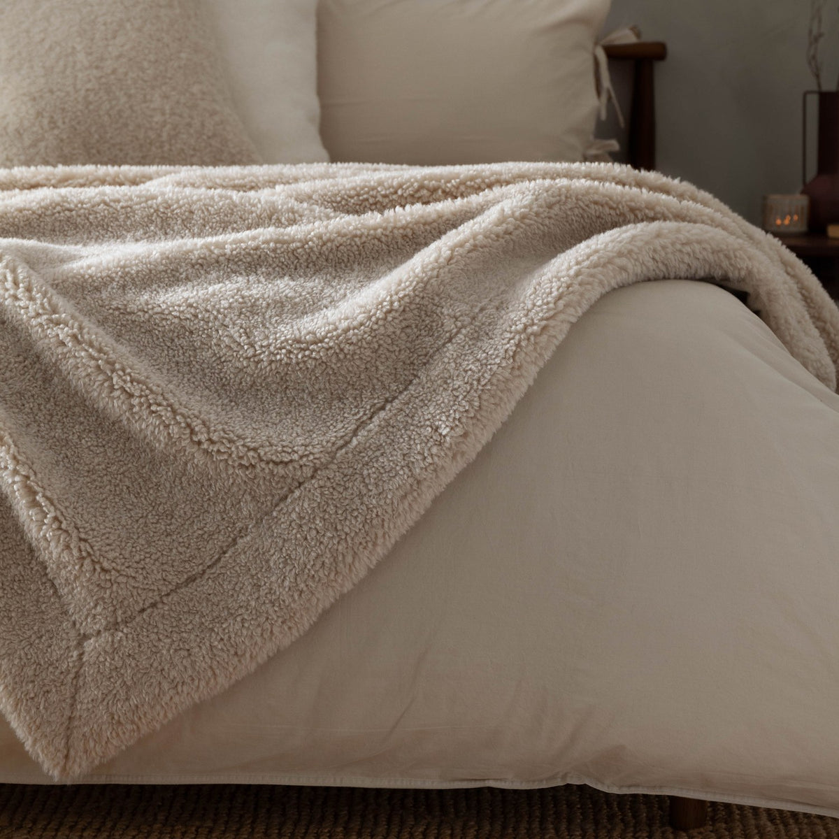 Olann Plain Velvet Throw in Natural, draped over a bed showing its shearling-look faux fleece front and smooth velvet reverse, with pillows and headboard in the background.